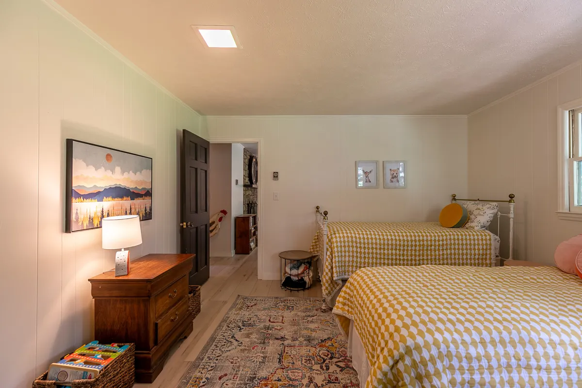 A cozy bedroom with two beds adorned in cheerful yellow checkered bedding, a wooden nightstand with a lamp, colorful area rug, and playful artwork, creating a warm space ideal for children or extra guests. The scene evokes the joy of shared sleepovers and story time.