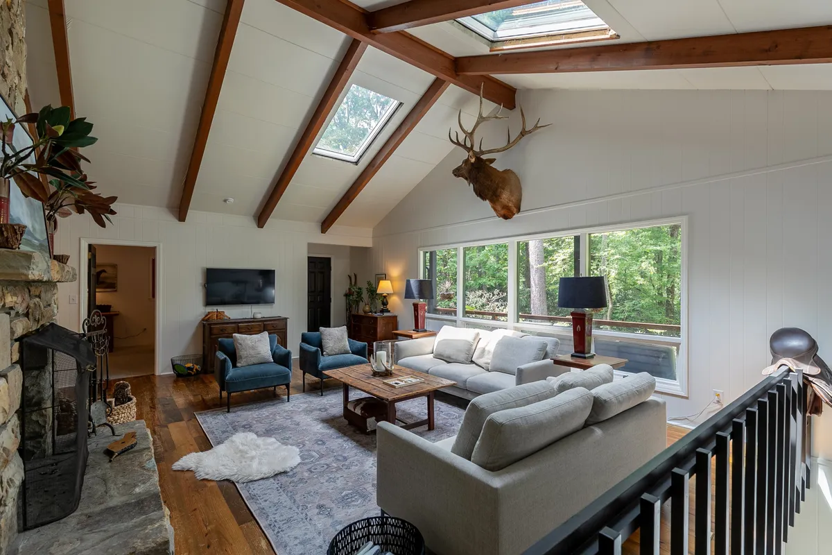 A spacious, sunlit living area featuring large windows with views of nature, a striking stone fireplace, comfortable seating, antler mount, and rich wooden beams. The scene invites group gatherings, whether for relaxing, entertaining, or enjoying the scenic outdoors through the windows.