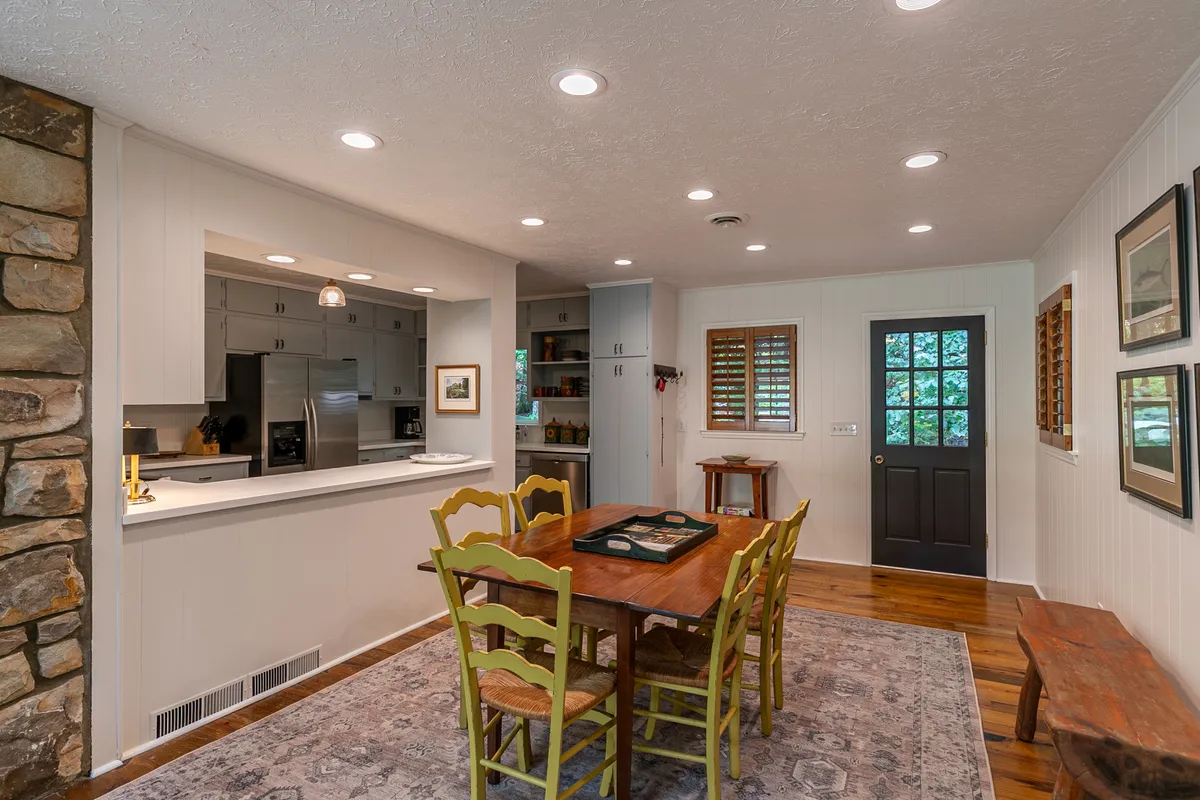 A cozy dining space with a stone accent wall, centrally located wooden dining table set for six, and an adjacent kitchen area. The space is bathed in soft natural light from surrounding windows, creating an inviting setting for shared meals or game nights.