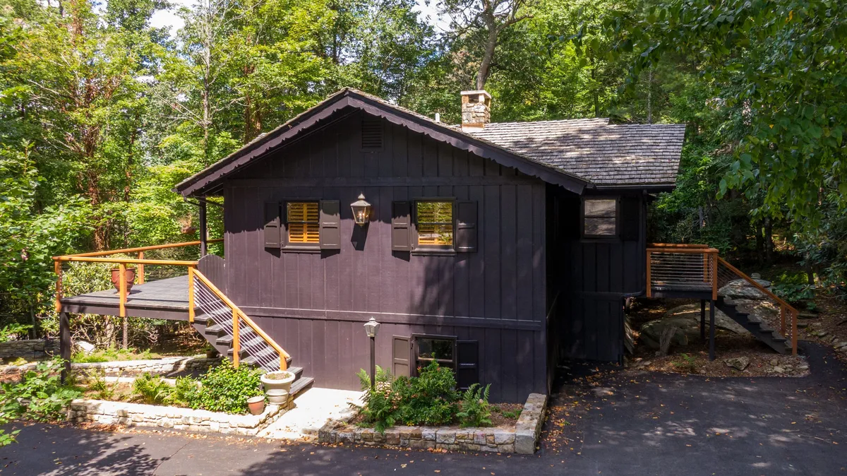 This image showcases the cabin’s dark wooden exterior, a stone pathway leading up to the deck, and lush greenery in the background. The deck is visible with seating, and the scene is bathed in warm light, creating an inviting atmosphere. There is a sense of tranquility, with hints of sunset and cozy outdoor moments, and the firepit area is also visible, suggesting gatherings after dark.