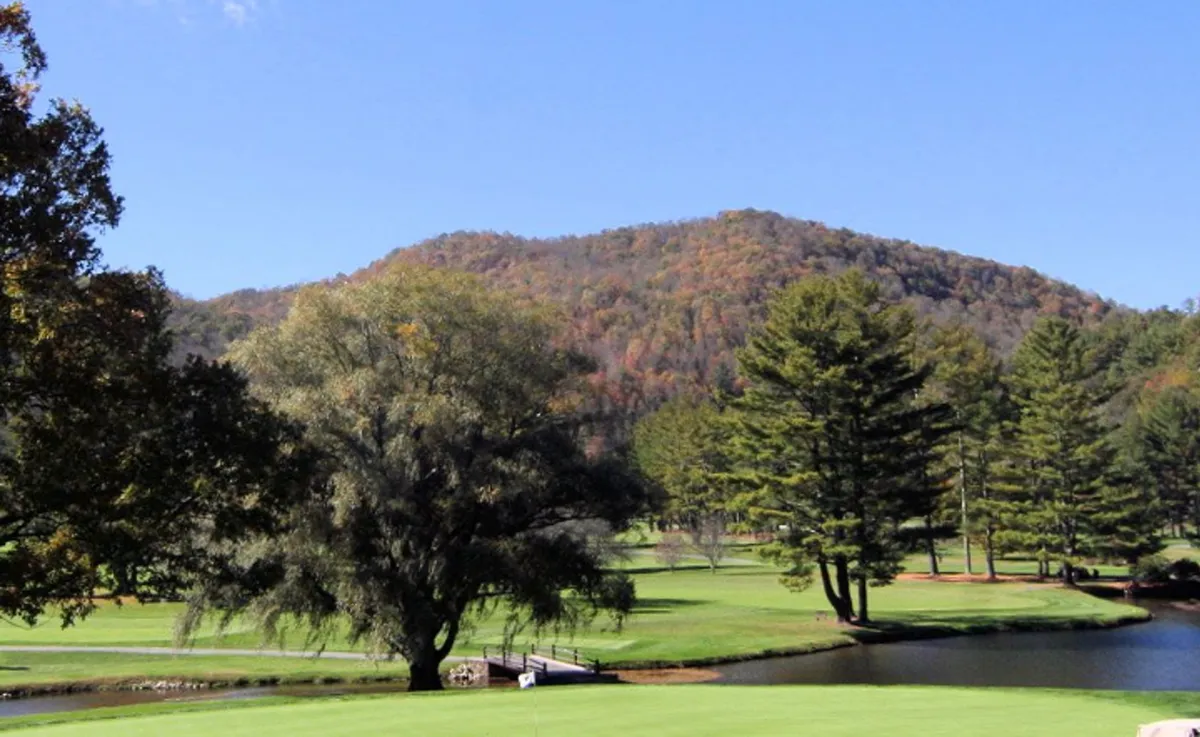 A scenic mountain view with a large tree in the foreground, lush golf course greenery, a tranquil pond, and the distant silhouette of mountains under a serene sky. The landscape exudes peacefulness and offers a sense of open space and natural beauty.