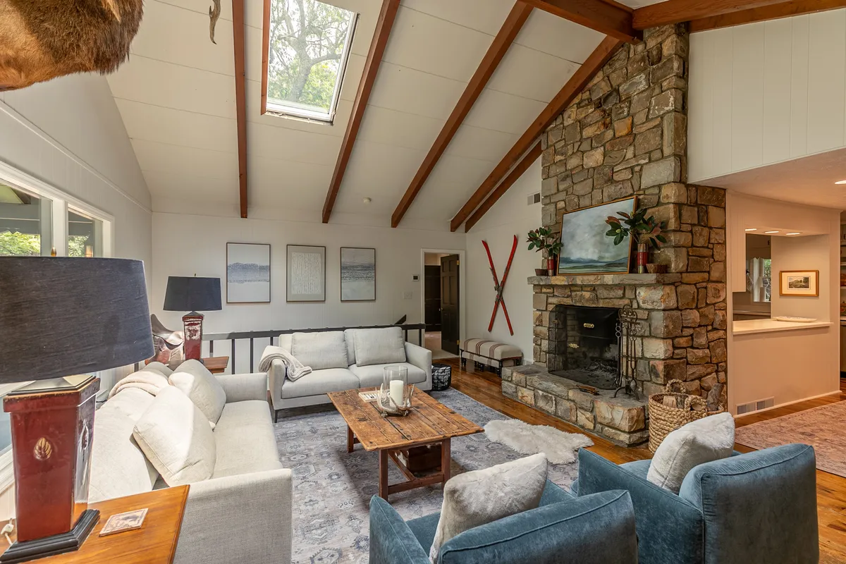 A warm, welcoming great room centered around a striking stone fireplace, accented by exposed wooden beams and a plush sofa set. Large windows allow sunlight to fill the space, and a coffee table invites gathering for games or conversation. The interior design exudes comfort and rustic charm.