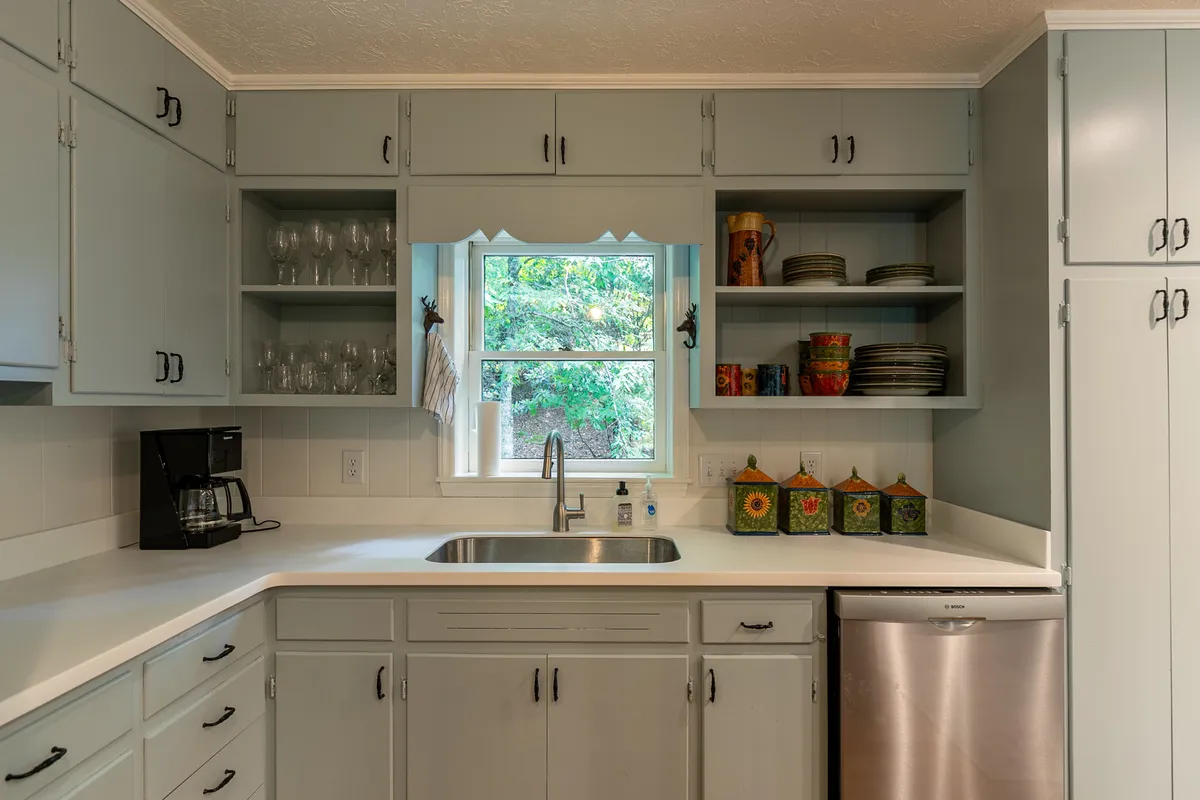 A bright kitchen scene features a sink positioned beneath a window that looks out onto lush greenery. The kitchen cabinets are warm-toned and inviting, with colorful dishware adding a lively touch. Natural light pours through the window, offering a tangible connection to the outdoors.