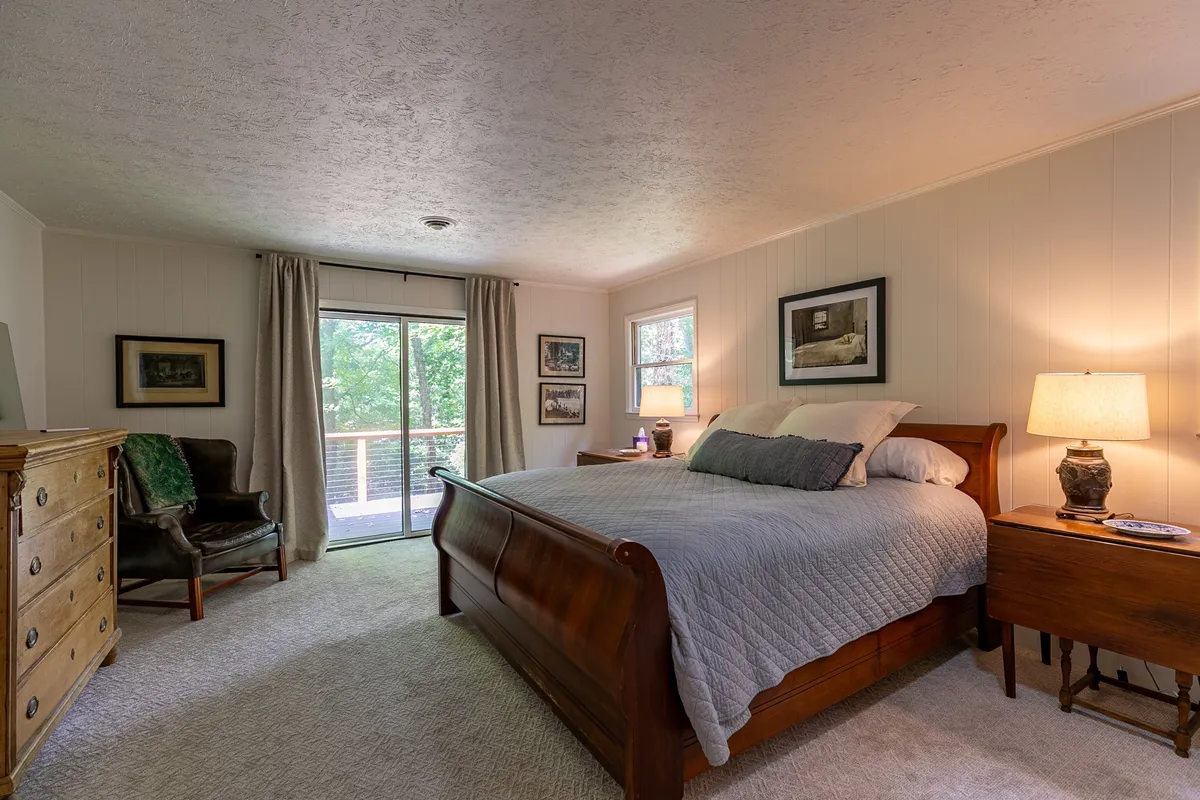 This image displays a serene bedroom with a sleigh bed, wooden dresser, and armchair. The focal point is the sliding glass door, which provides a visual and physical connection to the outdoors and hints at scenic nature views just beyond. Soft natural light fills the room, inviting a sense of tranquility.