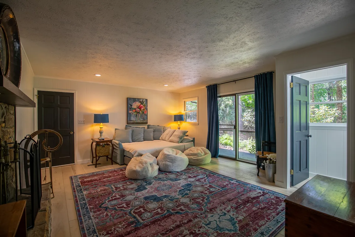 A den-like living area featuring a comfortable sofa, colorful rug, bean bags, and artwork, creating a welcoming environment for relaxing, reading, or socializing. The large windows suggest a connection to nature and ample natural light.