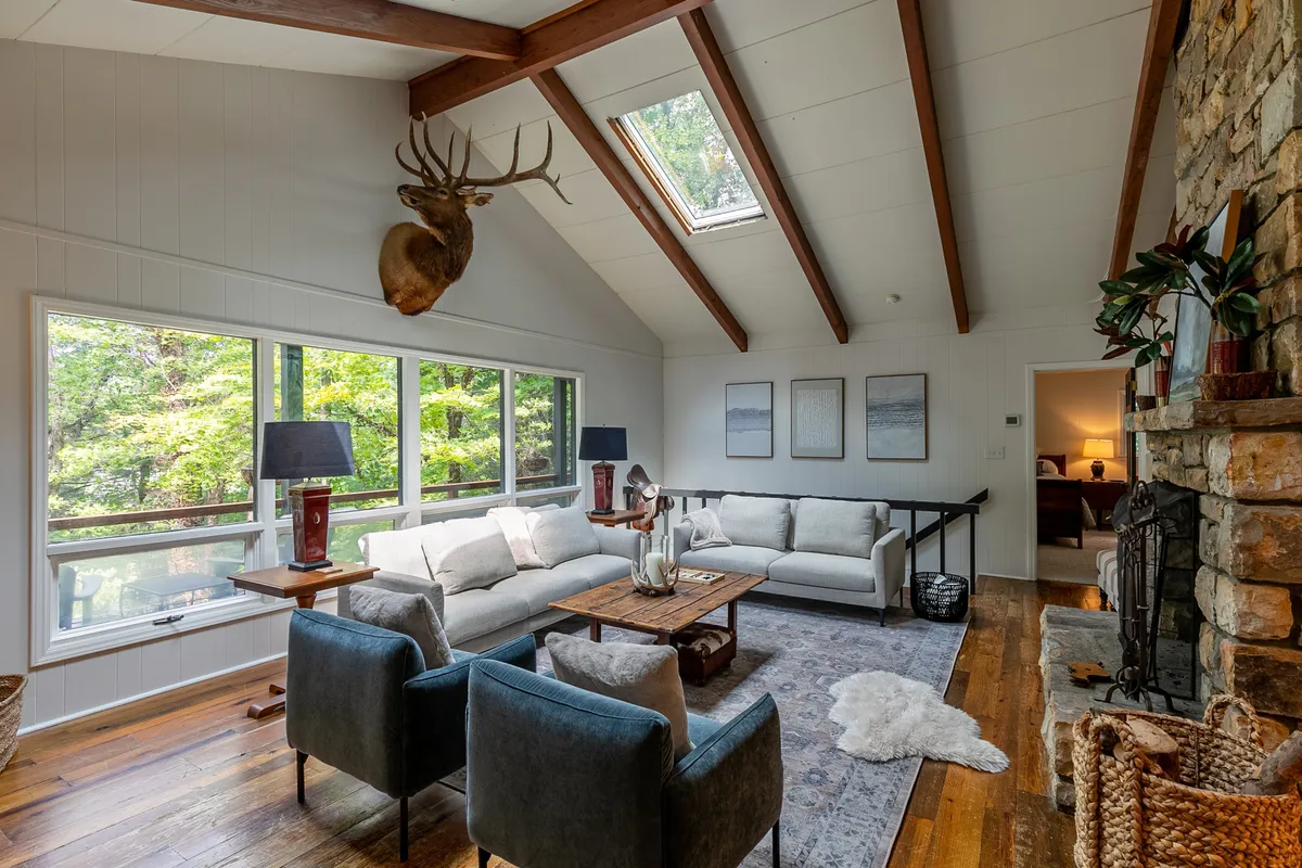 A warm, inviting living space featuring large windows overlooking lush greenery, a cozy stone fireplace, a comfortable sofa set, and decorative antlers mounted on the wall. The room is brightened by natural light and exudes a welcoming, family-friendly atmosphere.