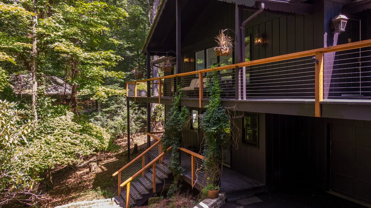 A modern wooden deck surrounded by lush green foliage, with stylish railing and exterior lighting. The space invites guests to sit, relax, and take in the surrounding natural beauty. The covered and open-air seating options are implied, and the environment is serene and welcoming, ideal for gatherings and outdoor enjoyment.