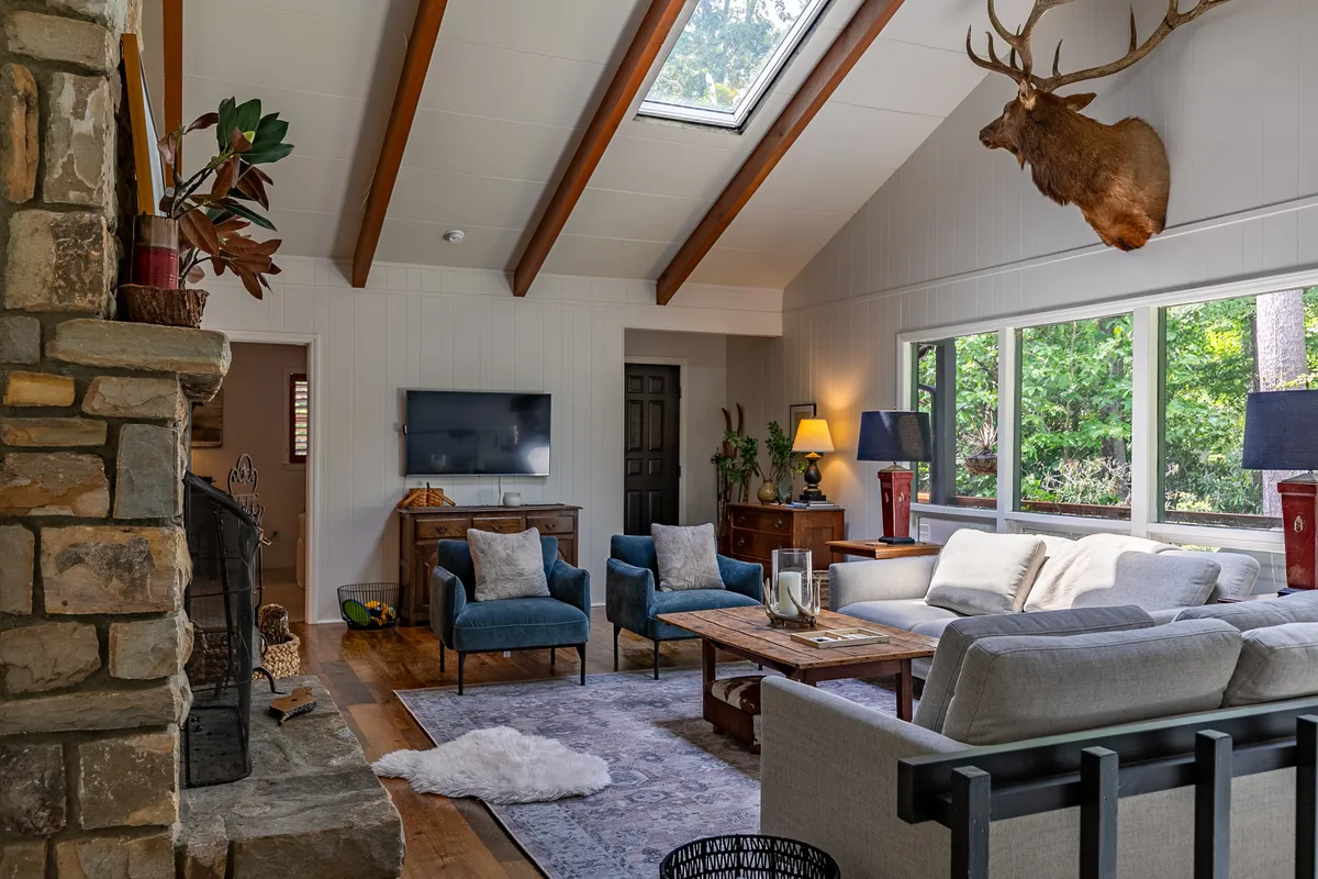 A welcoming living area centered around a grand stone fireplace, with a plush sofa, large windows that flood the space with natural light, and a mounted deer head lending a rustic, mountain-lodge ambiance. The wooden coffee table and comfortable seating create an ideal spot for gathering after a day of adventure.